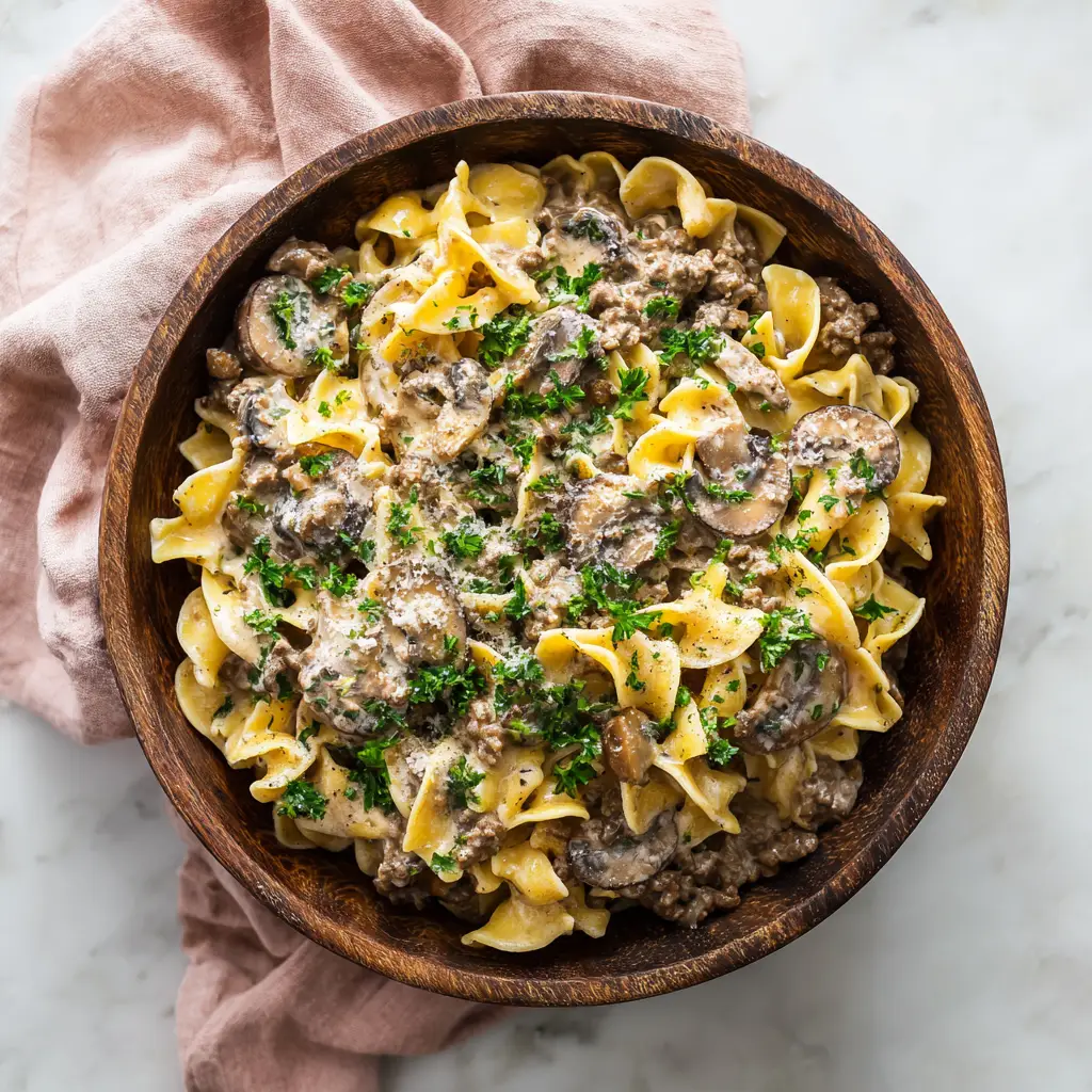 Ground Beef Stroganoff: A 30-Minute Rustic Masterpiece 1 An overhead shot of the finished Ground Beef Stroganoff in a dark wooden bowl, set on a white marble surface next to a soft pink napkin, emphasizing the dish's beautiful presentation.
