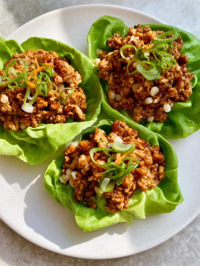 PF Chang’s Chicken Lettuce Wraps: The Ultimate 20-Minute Copycat 2 A close-up top-down view of three vibrant green butter lettuce cups filled with browned minced chicken and a dark hoisin glaze on a white ceramic plate. (PF Chang’s Chicken Lettuce Wraps)