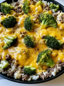 Angled view of a black cast iron skillet filled with glossy Cheesy Ground Beef and Broccoli Rice, showing cracked pepper and dried herbs. (Cheesy Ground Beef and Broccoli Rice)