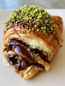 A handheld photo of a golden-brown flaky Pistachio Chocolate Croissant showing buttery pastry layers.
